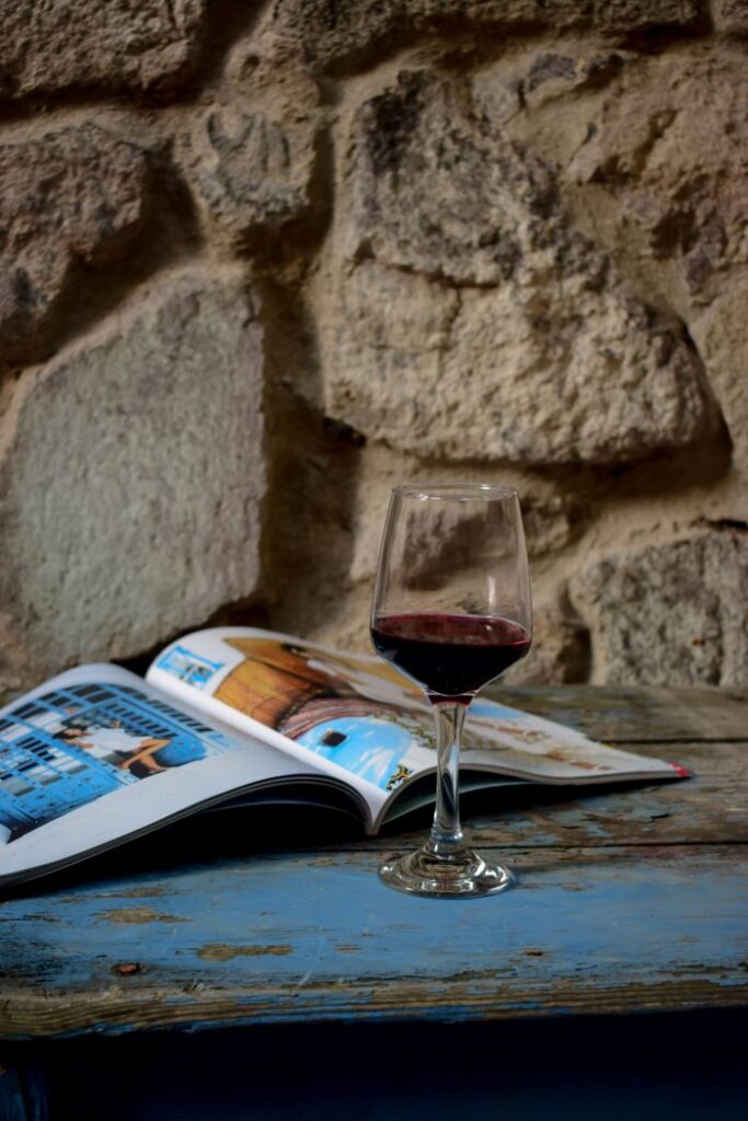 A glass of red wine on a wooden table beside an open travel magazine against a stone wall.