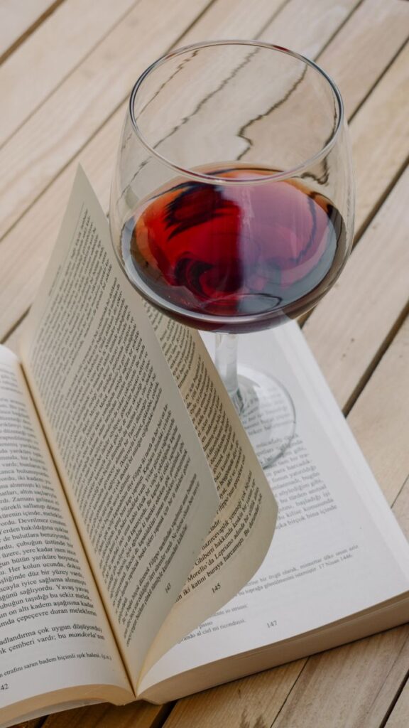 A glass of red wine rests on an open book on a wooden table, creating a cozy reading scene.